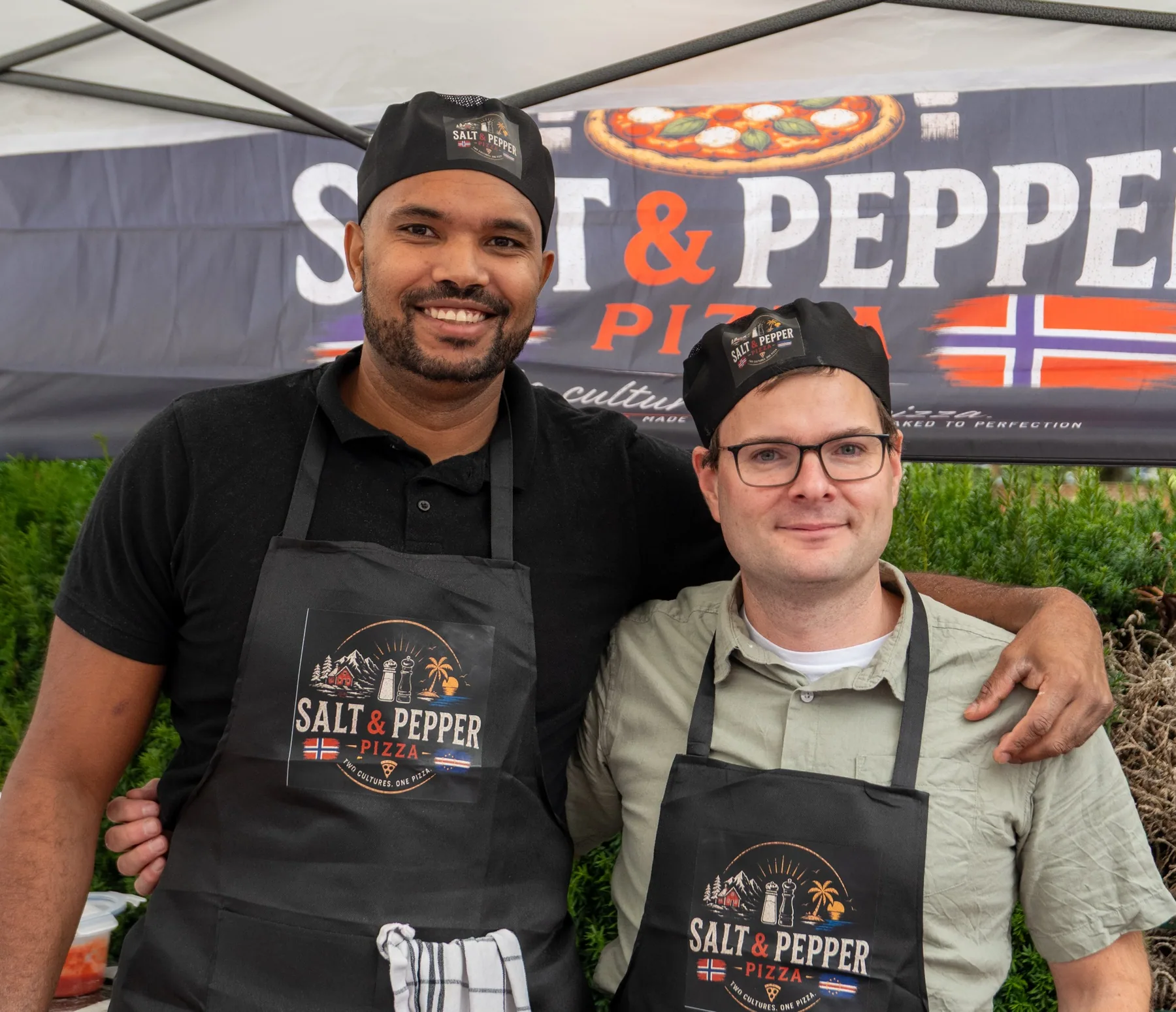 To deltakere i Salt & Pepper Pizza-forklær foran markedsbanneret